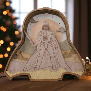 May include: A wooden, layered art piece featuring Darth Vader holding a lightsaber. The artwork is in the shape of Darth Vader's helmet, with a landscape and a sun in the background. The piece is made of light-colored wood.