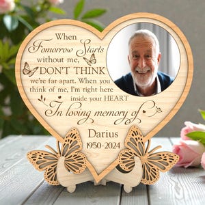 Può includere: Una cornice portafoto commemorativa a forma di cuore in legno con la foto di un uomo. La cornice presenta il testo "When Tomorrow Starts without me, DON'T THINK...In loving memory of Darius 1950-2024" e ritagli di farfalle. La cornice è sostenuta da due gambe a forma di farfalla.