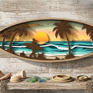 Puede incluir: Una pieza de arte mural de madera con forma de tabla de surf que representa una escena de playa con un surfista, palmeras y una puesta de sol. La obra de arte está en capas, creando un efecto 3D. Conchas marinas, vidrio de mar y una pequeña planta en maceta se exhiben debajo.