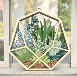 Może przedstawiać: Drewniane geometryczne terrarium z różnorodnymi, warstwowymi sukulentami. Terrarium ma ramę z naturalnego drewna i jest przeznaczone do zawieszenia. Sukulenty są w odcieniach zieleni, błękitu i bieli.