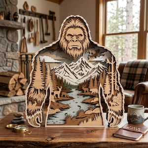 May include: A multi-layered wooden wall art piece shaped like a Bigfoot silhouette, featuring a detailed laser-cut landscape with mountains, pine trees, a river, and various forest animals including deer, a bear, and a fox in natural wood tones.