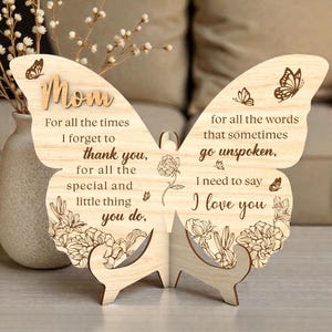 Op de afbeelding: Een houten vlindervormig ornament met het woord "Mom" bovenaan. De vleugels zijn versierd met gegraveerde tekst en bloemmotieven. De tekst luidt: "Voor alle keren dat ik vergeet je te bedanken, voor alle speciale en kleine dingen die je doet, ik hou van je."