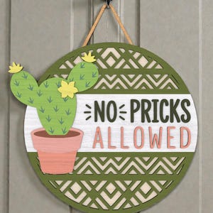May include: Round wooden sign with a green border and a white center. The sign features a green cactus in a pink pot and the text "NO PRICKS ALLOWED" in black and pink. A rope is attached for hanging.