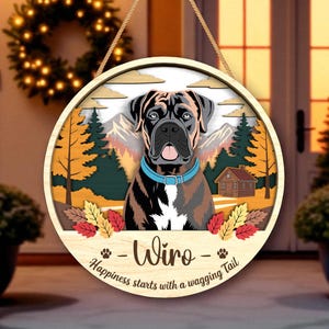 May include: A round wooden sign featuring a Boxer dog with a blue collar, set against a mountain and forest backdrop. The sign includes the name "Wiro" and the phrase "Happiness starts with a wagging tail."