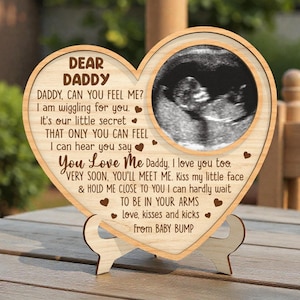 Op de afbeelding: Houten hartvormige plaquette met een echobeeld en de tekst "Lieve Papa". De plaquette bevat een standaard en heeft gegraveerde tekst die liefde en verwachting voor een baby uitdrukt. Het frame is gemaakt van lichtgekleurd hout.