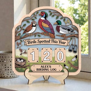 May include: Wooden birding log with a decorative design. The sign features illustrations of birds, a nest, binoculars, and feathers. The text "Birds Spotted This Year" and "Alex's Birding Log" are displayed. The counter shows the number 120.
