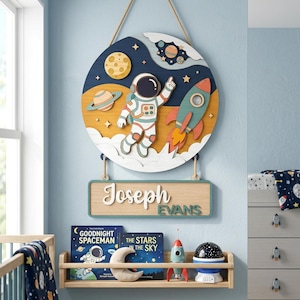 May include: A wooden space-themed wall hanging featuring an astronaut, rocket, and planets. The name "Joseph Evans" is displayed on a rectangular sign below. The design uses a blue, yellow, and white colour scheme, suitable for a child's room.