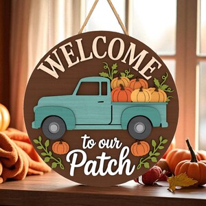 May include: A round wooden sign with the words "WELCOME to our Patch" and a teal pickup truck carrying pumpkins. The sign is brown with white lettering and a jute rope for hanging. Pumpkins and fall foliage are also visible.
