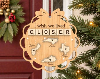 May include: Wooden Christmas ornament with the text "I wish we lived CLOSER." Features states with heart details and names. Includes a red and white ribbon for hanging. A festive holiday decoration.