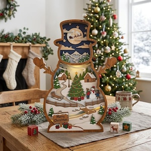 May include: A wooden snowman-shaped light box featuring a layered winter scene with a Christmas tree, houses, and a bridge. The top section shows a moonlit sky with a silhouette of Santa's sleigh. A sign at the bottom reads Merry Christmas.