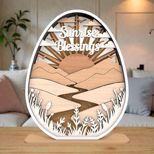 May include: Wooden layered art piece in an egg shape, featuring a sunrise scene with mountains, a river, and the text "Sunrise Blessings." The piece has a white frame and a wooden base, with intricate cut-out details.