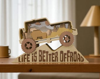 May include: A layered wooden sculpture of an off-road vehicle, featuring a mountain scene inside. The sculpture is mounted on a base with the text "LIFE IS BETTER OFFROAD" in brown. The wheels are a light brown color.