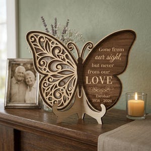 May include: A wooden butterfly-shaped memorial plaque with intricate cut-out designs. The right wing features the text "Gone from our sight, but never from our LOVE" and the name "Escobar 1958-2026". A framed photo and a lit candle are also present.