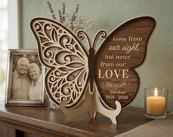 May include: A wooden butterfly-shaped memorial plaque with intricate cut-out designs. The right wing features the text "Gone from our sight, but never from our LOVE" and the name "Escobar 1958-2026". A framed photo and a lit candle are also present.