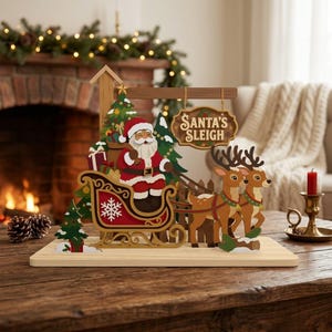 Op de afbeelding: Een houten tafeldecoratie met een gelaagd uitgesneden ontwerp van de Kerstman in een rode slee getrokken door twee rendieren. De scène bevat groene dennenbomen, ingepakte cadeautjes en een hangend bord met de tekst SANTA'S SLEIGH in bruine letters.