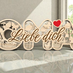 Op de afbeelding: Houten bord met de tekst "OMA Liebe dich" en een rood hart. De letters zijn lichtbruin met uitgesneden bloemen- en vlindermotieven. De woorden "Liebe dich" zijn in een sierlijk lettertype geschreven, wat een decoratief tintje geeft.