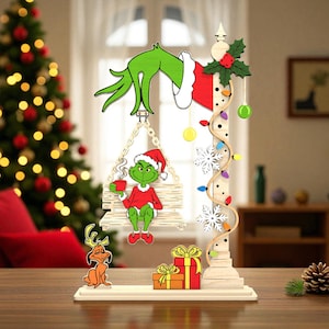 May include: A festive wooden Christmas ornament. It depicts a green character in a Santa hat on a swing, holding a mug. The design includes fairy lights, snowflakes, and a small dog figure. Presents are at the base of the ornament.
