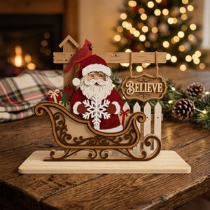 May include: A wooden tabletop decoration featuring a Santa Claus figure in a sleigh, holding a white snowflake. A red cardinal sits on a wooden fence post behind him, and a hanging sign with the word BELIEVE is suspended by twine.
