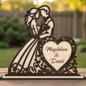 Puede incluir: Adorno de pastel de boda de madera con una pareja abrazándose. El diseño incluye un corazón con los nombres "Magdalene & David" grabados. Hecho de madera marrón clara y oscura, es un elemento decorativo.