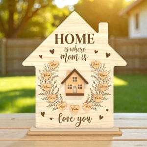 May include: Wooden house-shaped sign with the text "HOME is where mom is love you". The sign has a small house cutout in the centre and heart-shaped name tags. The sign is made of light-coloured wood.