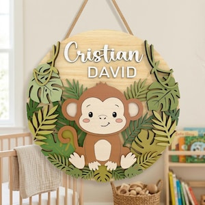 Op de afbeelding: Een ronde houten plaat met een lachende cartoon aap omringd door groene en bruine bladeren. De plaat heeft de naam "Cristian DAVID" in witte letters. Het hangt aan een touw.