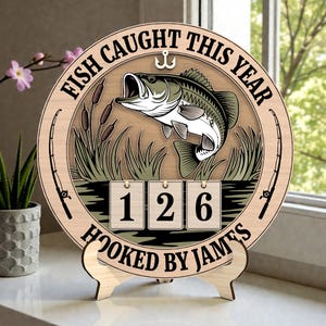 Puede incluir: Un letrero de madera con temática de pesca con el texto "FISH CAUGHT THIS YEAR" y "HOOKED BY JAMES". El letrero presenta una ilustración de un pez, cañas de pescar y un contador que muestra "126". El letrero se muestra en un soporte de madera.