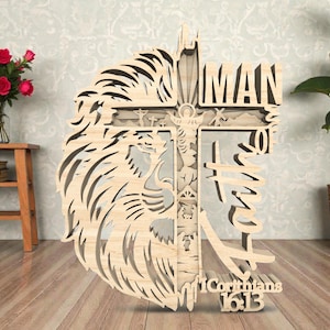 May include: Wooden wall art featuring a lion's head and a cross design. The cross includes the word "MAN" and the text "I Corinthians 16:13". The artwork is made of light-colored wood and has a layered design.