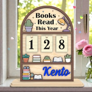 May include: Wooden book counter with the text "Books Read This Year" and the number "128". The counter has a brown frame with a light wood background and blue text that says "Kento". Decorative elements include a cactus, a lamp, and small books.