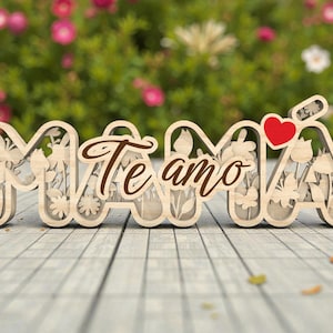 Op de afbeelding: Houten "MAMA Te amo" bord met een hart. De letters zijn lichtgekleurd met bloemen en vlinder uitsparingen. De woorden "Te amo" staan in een cursief lettertype. Een rood hart staat boven de letter "A". Het bord staat op een houten oppervlak.