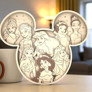 May include: A wooden, layered art piece in the shape of Mickey Mouse ears, featuring illustrations of Disney princesses. The princesses are depicted in a light brown color, with a light brown border. The artwork is displayed on a light-colored surface.