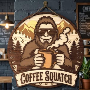May include: A circular wooden wall sign featuring a layered cutout design of a Sasquatch wearing sunglasses and holding a steaming mug of coffee. The background depicts mountains and pine trees. The text COFFEE SQUATCH is engraved at the bottom.