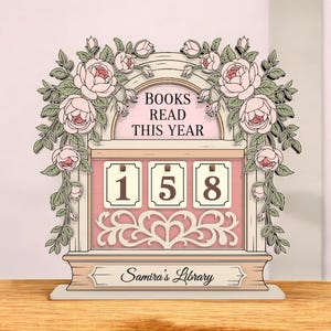May include: A decorative wooden book counter with the text "BOOKS READ THIS YEAR" and the number "158". The counter is adorned with pink roses and the words "Samira's Library".