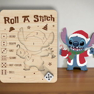 Op de afbeelding: Houten "Roll A Stitch" spelbord met een dobbelsteen en een Stitch-figuur. Het bord heeft uitsparingen voor Stitch's lichaamsdelen en instructies. Een aparte Stitch-figuur in een kerstmanoutfit is ook aanwezig, met een beker en een bel.
