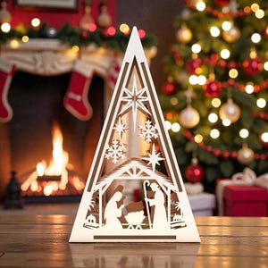 May include: A white, laser-cut wooden Christmas tree decoration depicting a nativity scene. The tree-shaped ornament has star and snowflake cutouts. The background includes a fireplace, Christmas tree, and presents.