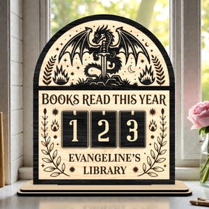 Op de afbeelding: Houten boekensteller met een draak- en zwaardontwerp. De teller leest "BOOKS READ THIS YEAR" en "EVANGELINE'S LIBRARY". De cijfers 1, 2 en 3 worden weergegeven. De teller is gemaakt van licht en donker hout.