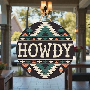 May include: Round wooden sign with a black background and the word "HOWDY" in large, white letters. The sign features a Southwestern-inspired design with geometric patterns in teal, orange, and white. A jute rope is attached for hanging.