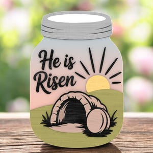 May include: A wooden Easter decoration in the shape of a mason jar. The jar features the text "He is Risen" in black script, a sunrise, and a depiction of the empty tomb. The color palette includes pastel pink, green, and yellow.