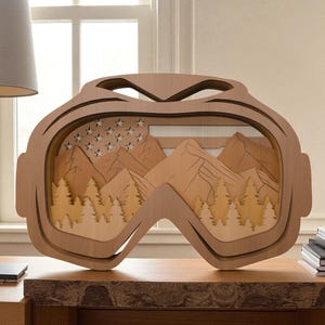 May include: A wooden art piece shaped like ski goggles, featuring a layered design of mountains, trees, and stars. The artwork is made of light brown wood and sits on a wooden surface.