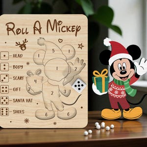 Op de afbeelding: Houten "Roll A Mickey" spelbord met een Mickey Mouse-uitsparing en dobbelstenen. Het bord heeft uitsparingen voor Mickey's lichaamsdelen, een dobbelsteen en de tekst "Roll A Mickey". Een Mickey Mouse-figuur met een kerstmuts staat naast het bord.