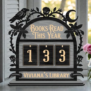 Op de afbeelding: Zwarte en grijze houten boekensteller met de tekst "Books Read This Year" en het nummer "133". Het ontwerp bevat een raaf, rozen en een halve maan. De tekst "Viviana's Library" staat onderaan.