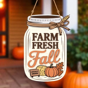 May include: Wooden mason jar-shaped sign with the words "Farm Fresh Fall" in brown and orange. The sign features a pumpkin, hay bale, and wheat stalk. A small sign reads "Locally Grown". The sign hangs from a brown string.