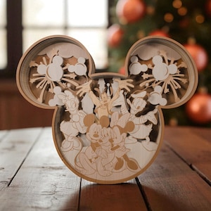 Könnte beinhalten: Ein Holz-Kunstwerk in Form von Mickey Mouse. Das Kunstwerk zeigt Mickey, Minnie, Donald, Daisy und Goofy. Das Design umfasst geschichtete Ausschnitte der Charaktere und Feuerwerk. Das Stück steht auf einem Holztisch mit einem Weihnachtsbaum im Hintergrund.