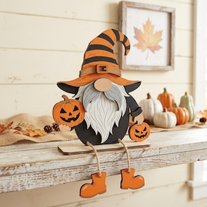 May include: A layered wooden gnome decoration featuring a tall orange and black striped hat, a long gray beard, and a black body. The gnome holds two small orange jack-o'-lanterns and has dangling legs with orange boots attached to a flat base.