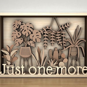 Puede incluir: Obra de arte enmarcada en madera con un diseño en capas de plantas en macetas, incluyendo monstera y suculentas. La frase "Just one more" está recortada en la parte inferior. La obra es en tonos marrones, con acabado de madera natural.
