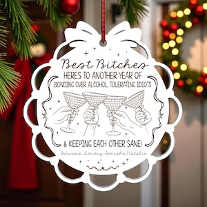 May include: White, laser-cut Christmas ornament with the text "Best Bitches" and a humorous quote about friendship. The ornament features a scalloped edge and a red ribbon for hanging. A festive holiday decoration.