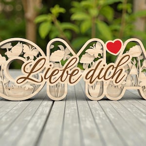 Pode incluir: Placa decorativa de madeira com a palavra "OMA" com recortes florais e de borboletas, e um coração vermelho. As palavras "Liebe dich" estão escritas em um script marrom na placa. A placa está em uma superfície de madeira cinza.