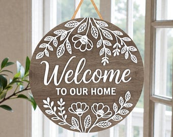 Könnte beinhalten: Runder Holz-Willkommensschild mit weißen Blumen- und Blattausschnitten. Auf dem Schild steht "Welcome TO OUR HOME" in weißer Schrift. Zum Aufhängen wird ein beiges Seil verwendet.