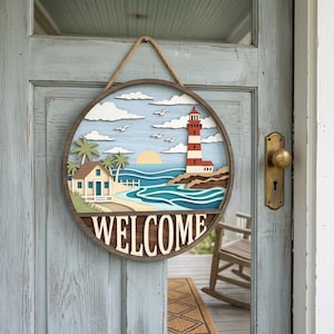 Può includere: Un cartello di benvenuto rotondo in legno con una scena costiera. Il cartello presenta un faro, spiaggia, oceano e la parola "WELCOME" in lettere bianche. È appeso a una corda di iuta ed è montato su una porta blu.