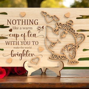Puede incluir: Letrero de madera con la cita "Nothing like a warm cup of tea with you to make the world brighter". Presenta un diseño de tetera y detalles de corazón. El letrero está sostenido por un soporte de madera y se exhibe sobre una superficie de madera.