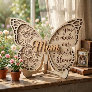Op de afbeelding: Houten vlindervormige decoratie met het woord "Mom" in het midden. De vlindervleugels hebben bloemmotieven en de woorden "you make our world bloom". De decoratie is lichtbruin en staat op een houten oppervlak.